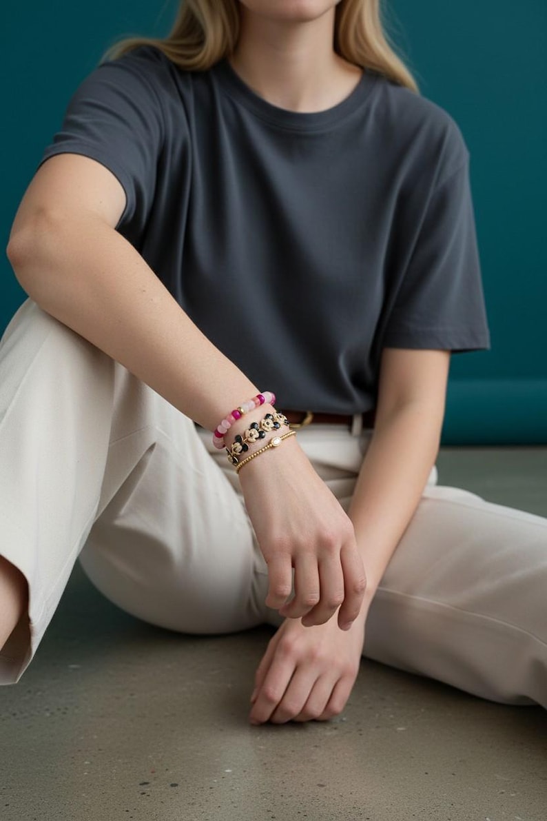 May include: A person wearing a dark gray t-shirt and light beige pants. The person is wearing three bracelets: one with pink beads, one with black flower-shaped beads, and a gold bracelet with a pearl.