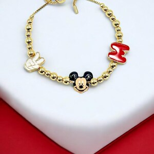 May include: Gold-toned beaded bracelet featuring Mickey Mouse, the peace sign, and the letter "M" in red enamel. The bracelet has a sliding clasp for adjustable sizing. The Mickey Mouse charm is black, white, and yellow.