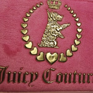May include: Close-up of a pink, plush fabric with a gold-colored emblem. The emblem features a dog, a crown, and heart-shaped accents, surrounded by a heart-shaped border. The words "Juicy Couture" are displayed in gold.