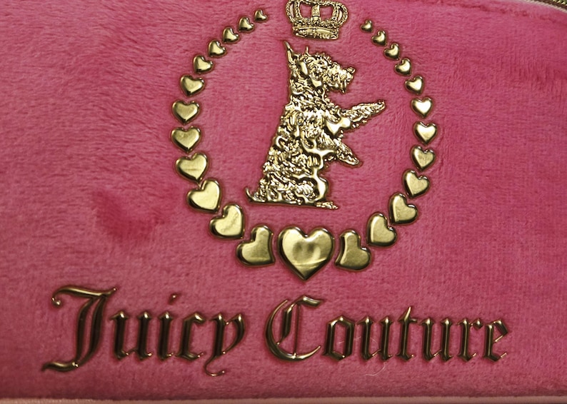 May include: Close-up of a pink, plush fabric with a gold-colored emblem. The emblem features a dog, a crown, and heart-shaped accents, surrounded by a heart-shaped border. The words "Juicy Couture" are displayed in gold.