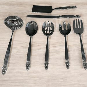 Könnte beinhalten: Ein Set silberfarbener Servierutensilien, darunter eine Kelle, ein Servierlöffel, ein geschlitzter Löffel, eine Serviergabel, ein Messer und ein Tortenheber. Jedes Teil hat einen dekorativen Griff.