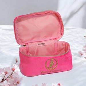 May include: Open pink velour cosmetic bag with a gold zipper and a gold emblem featuring a crown and the words "Juicy Couture". The interior is lined with pink fabric. The bag is open, revealing the inside.
