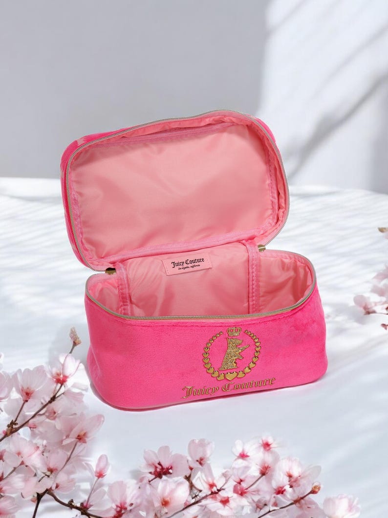 May include: Open pink velour cosmetic bag with a gold zipper and a gold emblem featuring a crown and the words "Juicy Couture". The interior is lined with pink fabric. The bag is open, revealing the inside.