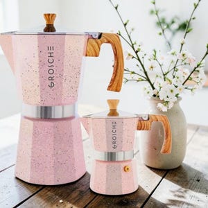May include: Two pink stovetop espresso makers with a speckled finish, silver metal bands, and wood-grain patterned handles and lid knobs. The brand name GROSCHE is printed vertically on the side of each pot. The pots have an octagonal base.