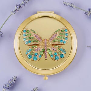 May include: A gold-colored, round compact mirror with a butterfly design. The butterfly is adorned with colorful rhinestones in shades of blue, green, and pink. The mirror is set against a lavender background, with sprigs of lavender on either side.