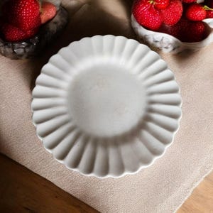 Może przedstawiać: Biały talerz z falistym brzegiem leży na beżowej lnianej powierzchni, obok dwóch małych misek wypełnionych czerwonymi truskawkami. Truskawki mają zielone łodygi i są zgrupowane.