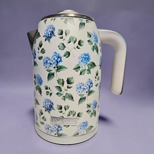 Può includere: Un bollitore elettrico color bianco sporco con un motivo ripetuto di fiori di ortensia blu e foglie verdi. Il bollitore presenta un coperchio argentato, un manico curvo, un beccuccio e una targhetta rettangolare argentata con la scritta Dwell Studio.