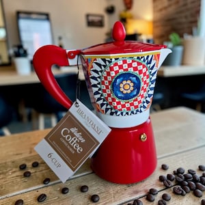 May include: A red 6-cup Italian coffee maker with a patterned top. The coffee maker has a red handle and a tag that reads "Italian Coffee Maker". Coffee beans are scattered around the coffee maker on a wooden surface.