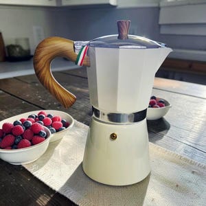 May include: A white moka pot with a wood-grain patterned handle and lid knob. The coffee maker features a metallic silver band around the center, a small gold-toned valve on the base, and a small tag with green, white, and red stripes near the handle.