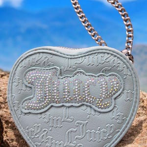 May include: A pale blue heart-shaped purse featuring the word "Juicy" in sparkling rhinestones. It has a silver chain handle and a repeating embossed "Juicy" pattern. The purse is a stylish accessory.