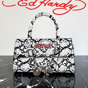 May include: A black and white snakeskin-effect handbag with a top handle. The bag has silver-coloured hardware and a chain with skull and rose charms. The red "Ed Hardy" logo is visible on the bag and above it.