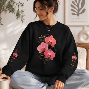 Peut inclure: Un sweat-shirt noir à manches longues orné d'un motif central représentant trois fleurs d'orchidée roses avec des tiges en lignes dorées délicates. Des motifs floraux d'orchidées roses assortis sont imprimés sur l'extérieur de chaque manche.