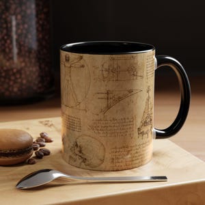 Peut inclure: Un mug en céramique avec un intérieur et une anse noirs, présentant un extérieur beige imprimé de croquis de Léonard de Vinci, incluant l'Homme de Vitruve, un crâne humain, des schémas mécaniques et des notes manuscrites à l'encre brune.