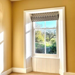 Könnte beinhalten: Ein Fenster mit weißem Rahmen und einem gemusterten römischen Rollo. Das Fenster blickt auf eine grüne Rasenfläche mit Bäumen im Hintergrund. Der Raum ist in einem hellen Gelb gestrichen.