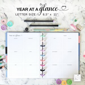 Könnte beinhalten: Ein Planer im Letter-Format mit dem Text "Year at a Glance" und einem Herzdesign. Der Planer ist geöffnet und zeigt monatliche Kalenderseiten. Das Bild enthält Stifte, dekoratives Klebeband und farbige Ringe. Der Planer misst 21,6 cm x 27,9 cm.