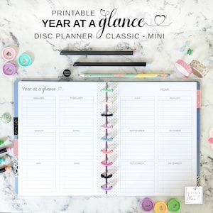 Könnte beinhalten: Ein druckbarer Year at a Glance Disc-Planer im klassischen Mini-Format. Der geöffnete Planer zeigt ein weißes Raster für zwölf Monate, das von bunten Kunststoffscheiben zusammengehalten wird. Enthalten sind zwei schwarze Stifte, ein gemusterter Bleistift und Klebebandrollen.