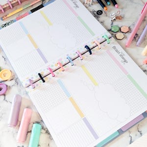 May include: A disc-bound planner open to a two-page spread with the text Brain dump. The pages feature pastel-colored grid and lined sections, a central cloud-shaped graphic, and a colorful disc spine. Surrounding the planner are markers, clips, and small geometric shapes on a white marble surface.