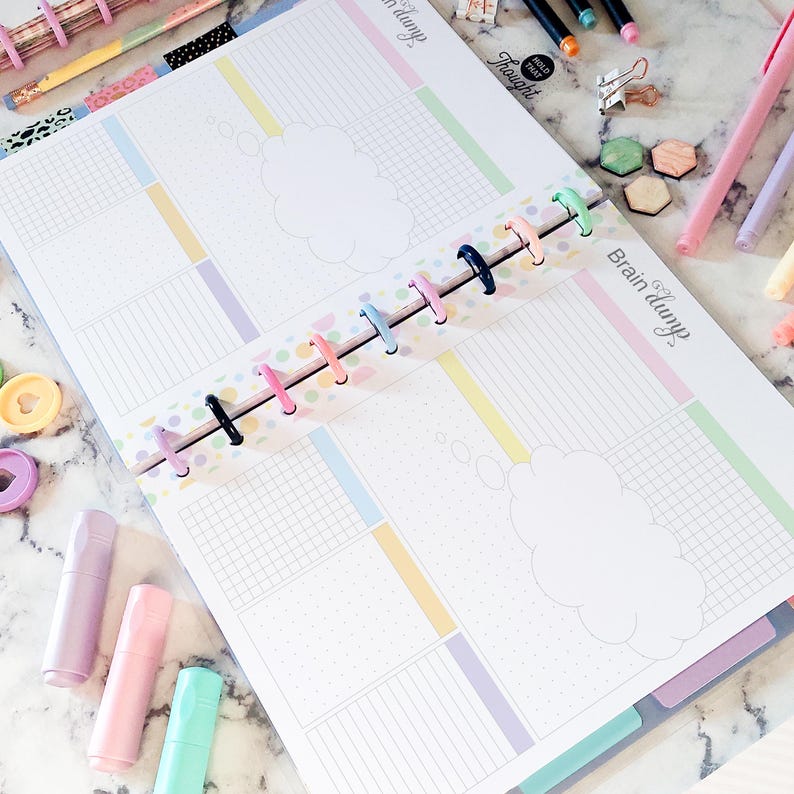 May include: A disc-bound planner open to a two-page spread with the text Brain dump. The pages feature pastel-colored grid and lined sections, a central cloud-shaped graphic, and a colorful disc spine. Surrounding the planner are markers, clips, and small geometric shapes on a white marble surface.