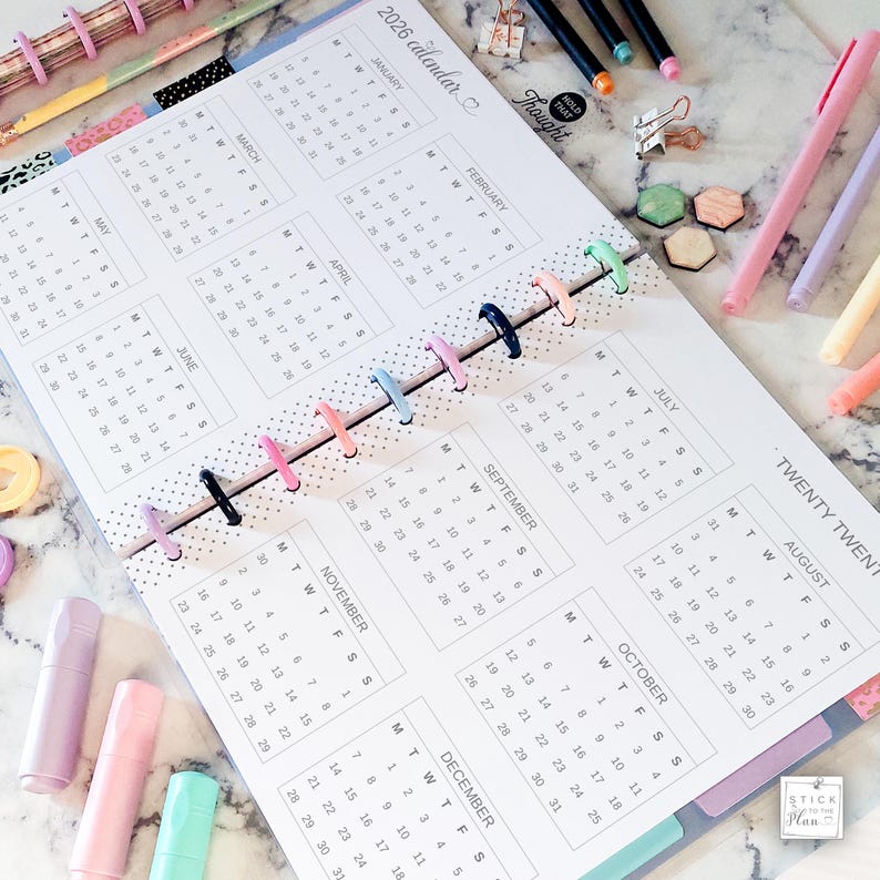 May include: A 2026 calendar planner with a white background and black text, featuring a disc-bound spine with colorful plastic rings. The layout displays all twelve months of the year, accompanied by various pastel-colored pens and small clips.