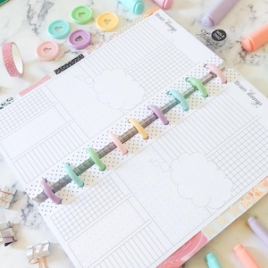 May include: Open planner with lined pages, thought bubbles, and the text "Brain dump." Colorful disc-bound rings, washi tape, and pens in pastel shades are scattered around the planner. Decorative heart-shaped discs are also visible.