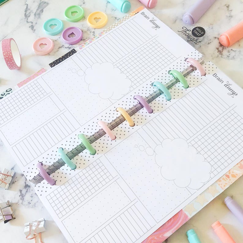 May include: Open planner with lined pages, thought bubbles, and the text "Brain dump." Colorful disc-bound rings, washi tape, and pens in pastel shades are scattered around the planner. Decorative heart-shaped discs are also visible.