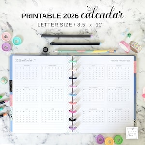 May include: A printable 2026 calendar with a letter size of 8.5" x 11". The calendar is open, showing the months of the year. There are pencils, washi tape, and decorative paper clips. The text "HOLD THAT Thought" is visible.
