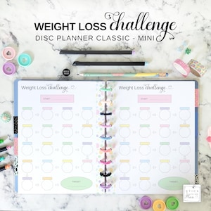 May include: A disc-bound planner open to a Weight Loss Challenge page with pastel-colored tracking boxes, arrows, and a target oval. The planner features a central spine with colorful discs, surrounded by two black pens, a pencil, and washi tape.