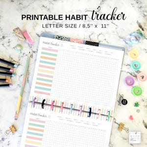 May include: A printable habit tracker with the text "PRINTABLE HABIT tracker LETTER SIZE / 8.5" x 11". The planner has a grid layout for tracking habits, with colorful dividers and a striped design. Includes washi tape, pencils, and binder clips.