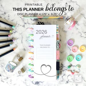 May include: A white 2026 planner with a heart design, colourful disc binding, and the text "This Planner Belongs To:". The image includes pencils, washi tape, binder clips, and decorative discs. The planner measures 10.48 cm x 23.5 cm.