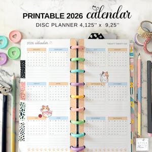 May include: A printable 2026 calendar disc planner, measuring 4.125" x 9.25". The calendar features monthly layouts with pastel color accents and cat illustrations. It is bound with colorful disc rings and includes decorative washi tape and pencils.