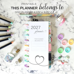 Op de afbeelding: Een 2027 disc planner van 10,48 cm bij 23,5 cm met een witte kaft, een hartafbeelding en de tekst THIS PLANNER BELONGS TO. De planner heeft kleurrijke schijven, zijtabbladen met patronen en is omringd door potloden, washi-tape, paperclips en losse plastic schijven.