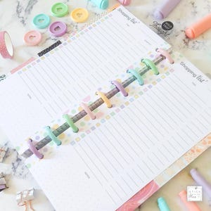 Puede incluir: Un planificador de lista de la compra de color pastel con una encuadernaci&oacute;n de anillos de colores. El planificador tiene un patr&oacute;n de puntos y p&aacute;ginas rayadas para escribir. El texto "Shopping List" est&aacute; impreso en la portada.