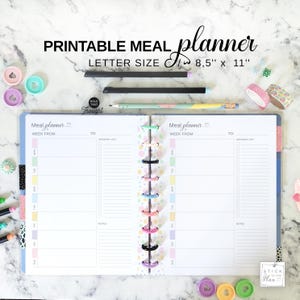 May include: Open printable meal planner with the text "PRINTABLE MEAL planner LETTER SIZE 8.5" x 11"" and "Meal planner". The planner is open to a blank page with a grocery list and notes section. The planner is held together with colorful rings.