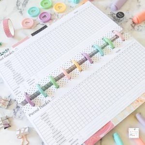 K&ouml;nnte beinhalten: Ein wei&szlig;er Planer mit einem Rastermuster und einem Abschnitt zur Verfolgung der Periode. Der Planer hat ein farbenfrohes Regenbogen-Design und die W&ouml;rter "Period tracker" und "Year" sind auf den Seiten gedruckt.