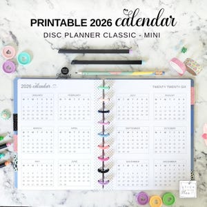 May include: A 2026 printable calendar for a disc planner classic mini, featuring a two-page spread with monthly grids from January to December. The planner includes colorful discs along the center binding, surrounded by pens and decorative washi tape.