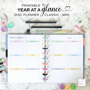 Könnte beinhalten: Ein druckbarer Jahresplaner mit Disc-Bindung im klassischen Mini-Format. Der geöffnete Planer zeigt ein Kalenderlayout für zwölf Monate mit pastellfarbenen Kopfzeilen für jeden Monat, zusammengehalten durch eine mittige Disc-Bindung.