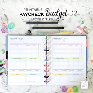 May include: A printable Paycheck Budget planner in letter size. The open planner shows a two-page spread with sections for income, bills, and expenses. The planner is held together with colorful rings and includes pens and decorative tape.