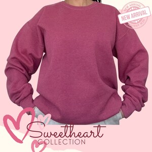 May include: A pink sweatshirt with a crew neck and long sleeves. The sweatshirt has a distressed look and is made of a soft, comfortable fabric. The text "Sweetheart Collection" is printed on the bottom of the sweatshirt.
