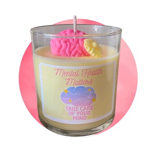 K&ouml;nnte beinhalten: Eine gelbe Kerze mit einem rosa Gehirn und einer gelben Blume oben drauf. Die Kerze hat ein Etikett mit der Aufschrift "Mental Health Matters" und "Take Care of Your Mind".