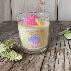 K&ouml;nnte beinhalten: Eine gelbe Kerze in einem Glasgef&auml;&szlig; mit einem rosa, gehirnf&ouml;rmigen Aufsatz. Das Etikett lautet "Mental Health Matters" und "Take Care of Your Mind".