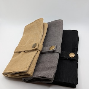 Könnte beinhalten: Drei Werkzeugrollen aus Canvas in Beige, Grau und Schwarz. Jede Rolle ist gefaltet und mit einem passenden Stoffriemen und einem messingfarbenen Druckknopfverschluss gesichert. Die Rollen sind nebeneinander vor weißem Hintergrund angeordnet.