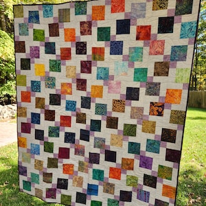 May include: A colorful handmade quilt with a repeating pattern of squares in various colors, including orange, green, purple, and blue. The squares are set against a neutral background and connected by lavender borders. The quilt is hanging outdoors on a clothesline.
