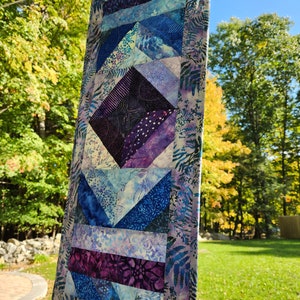 May include: A rectangular, quilted wall hanging with a geometric design. The quilt features shades of blue, purple, and white, with a central diamond shape and diagonal stripes. The edges are adorned with a fern pattern. It is hanging outside on a clothesline.
