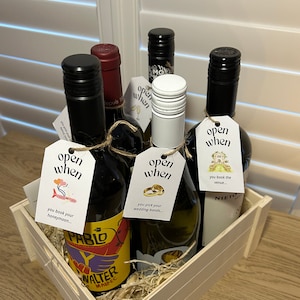 Puede incluir: Una caja de madera llena de seis botellas de vino, cada una con una etiqueta que dice "abrir cuando" y una ocasión diferente, como "reservas tu luna de miel" o "eliges tus alianzas".