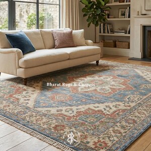 May include: A large rectangular area rug with a central floral design in shades of blue, beige, and red. The rug is placed in a living room setting with a beige sofa and a fireplace. The rug has a fringed edge.