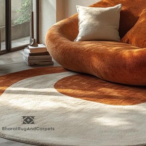 May include: A round, cream-colored rug with two brown, semi-circular patterns. The rug is in front of a brown, plush, modern sofa with white pillows. The text "BharatRugAndCarpets" is visible in the bottom left corner of the rug.