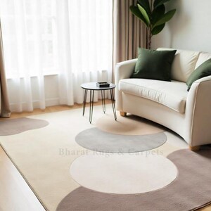 May include: A beige area rug with a geometric pattern of brown and gray semi-circles. The rug is in a living room setting with a white sofa and a black side table.