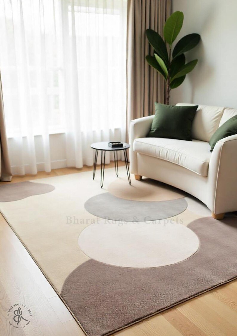 May include: A beige area rug with a geometric pattern of brown and gray semi-circles. The rug is in a living room setting with a white sofa and a black side table.