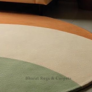 Puede incluir: Una alfombra redonda multicolor con tres rayas de color naranja, crema y verde. La alfombra tiene un brillo sutil y una textura suave. El texto "Bharat Rugs & Carpets" est&aacute; impreso en la alfombra.