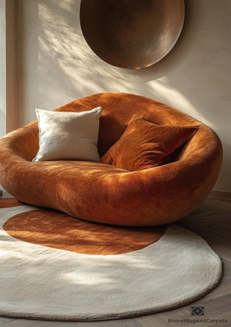 May include: A large, brown, plush, beanbag-style chair with two pillows. One pillow is white and the other is orange. The chair is sitting on a white and orange round rug.